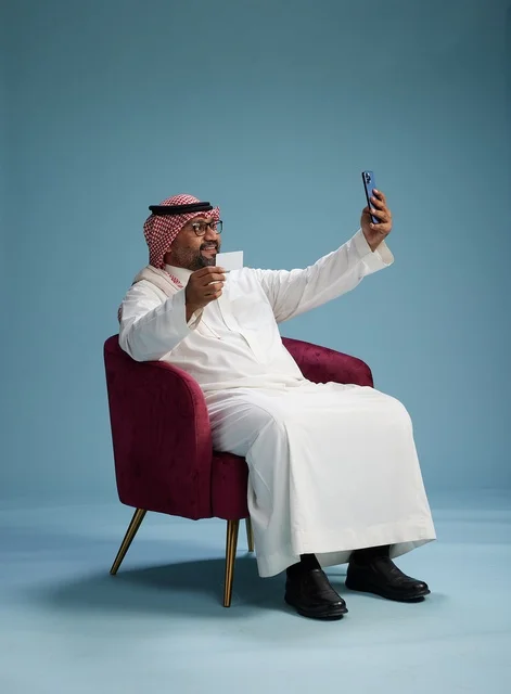 A portrait of a Saudi man using a mobile phone sitting on a chair while holding a blank white card, using a bank card, with a blue background.
