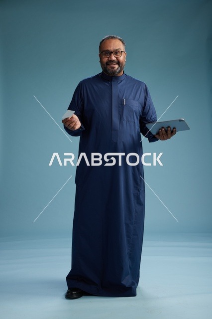 بورتريه لرجل سعودي يرتدي ثوباً أزرق يقرأ بطاقة عمل مستخدماً جهازاً لوحياً, استخدام البطاقه البنكيه , كامل الجسم  خلفيه زرقاء