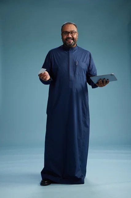 بورتريه لرجل سعودي يرتدي ثوباً أزرق يقرأ بطاقة عمل مستخدماً جهازاً لوحياً, استخدام البطاقه البنكيه , كامل الجسم  خلفيه زرقاء