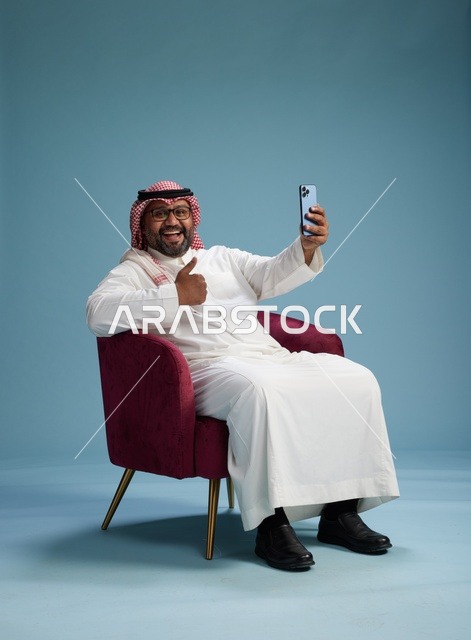بورتريه لرجل سعودي بالزي التقليدي يجلس ويلتقط سيلفي بالهاتف المحمول, خلفيه زرقاء