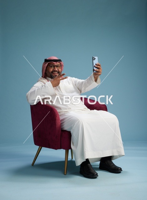 بورتريه لرجل سعودي بالزي التقليدي يجلس ويلتقط سيلفي بالهاتف المحمول, خلفيه زرقاء