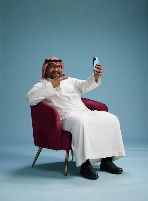 A portrait of a Saudi man in traditional attire sitting and taking a selfie with a mobile phone, with a blue background.