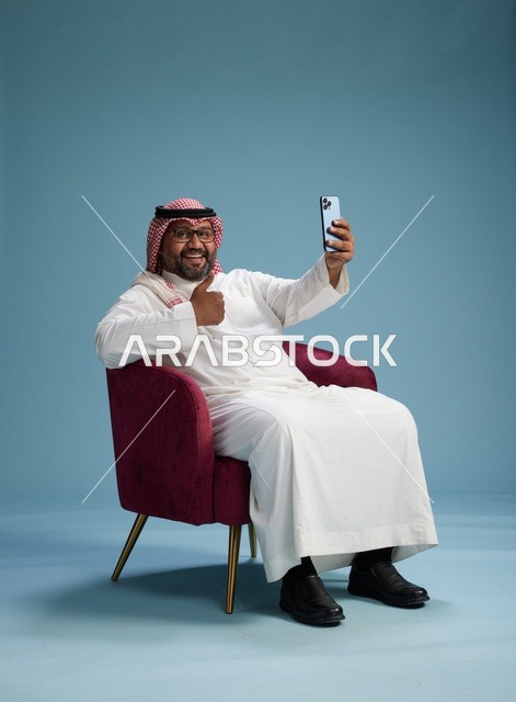 بورتريه لرجل سعودي بالزي التقليدي يجلس ويلتقط سيلفي بالهاتف المحمول, خلفيه زرقاء