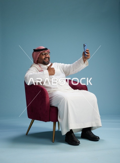 بورتريه لرجل سعودي بالزي التقليدي يجلس ويلتقط سيلفي بالهاتف المحمول, خلفيه زرقاء