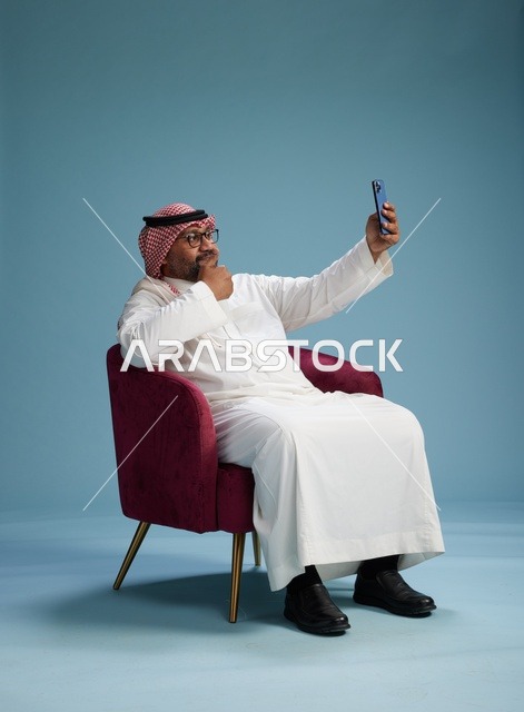 A portrait of a Saudi man in traditional attire sitting and taking a selfie with a mobile phone, with a blue background.