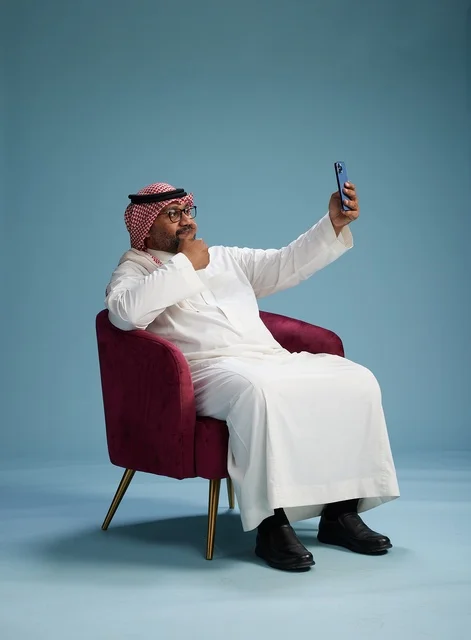 A portrait of a Saudi man in traditional attire sitting and taking a selfie with a mobile phone, with a blue background.