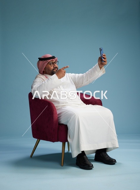 A portrait of a Saudi man in traditional attire sitting and taking a selfie with a mobile phone, with a blue background.