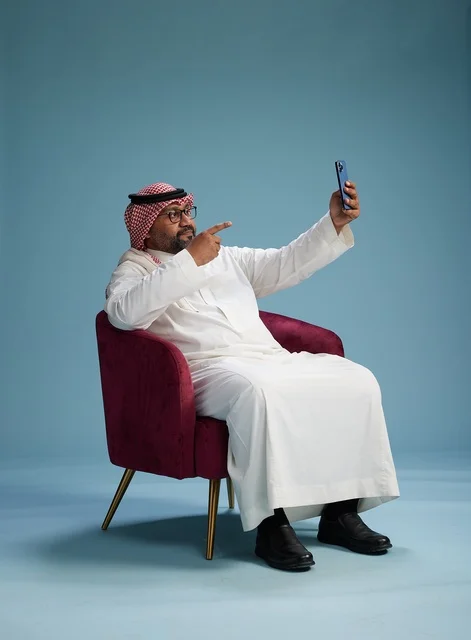 بورتريه لرجل سعودي بالزي التقليدي يجلس ويلتقط سيلفي بالهاتف المحمول, خلفيه زرقاء