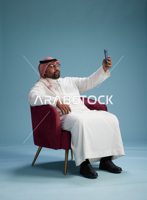 بورتريه لرجل سعودي بالزي التقليدي يجلس ويلتقط سيلفي بالهاتف المحمول, خلفيه زرقاء