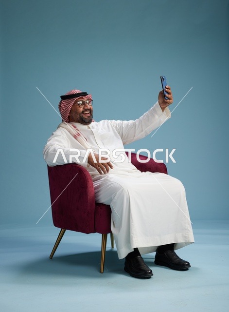 بورتريه لرجل سعودي بالزي التقليدي يجلس ويلتقط سيلفي بالهاتف المحمول, خلفيه زرقاء