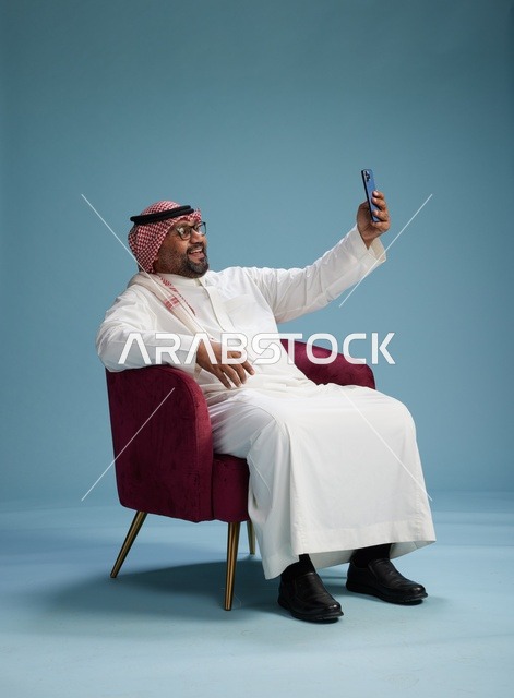 A portrait of a Saudi man in traditional attire sitting and taking a selfie with a mobile phone, with a blue background.