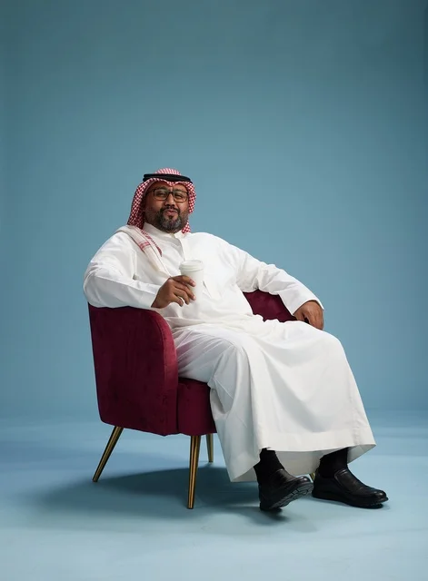 A portrait of an Arab Saudi Gulf man in a white thobe sitting holding a cup of coffee, enjoying hot beverages, with a blue background.