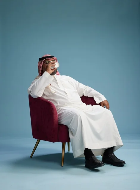 A portrait of an Arab Saudi Gulf man in a white thobe sitting holding a cup of coffee, enjoying hot beverages, with a blue background.