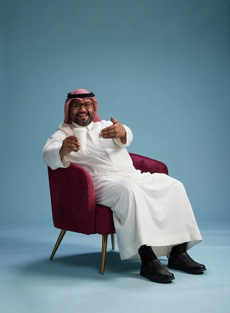 A portrait of an Arab Saudi Gulf man in a white thobe sitting holding a cup of coffee, enjoying hot beverages, with a blue background.