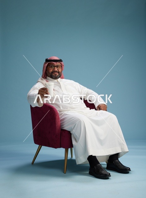 بورتريه لرجل عربي سعودي خليجي بثوب أبيض جالس ممسكاً بكوب قهوة, الاستمتاع بالمشروبات الساخنه, خلفيه زرقاء