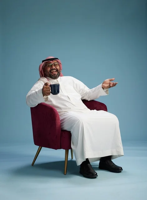A portrait of an Arab Saudi Gulf man in a white thobe sitting holding a cup of coffee, enjoying hot beverages, with a blue background.