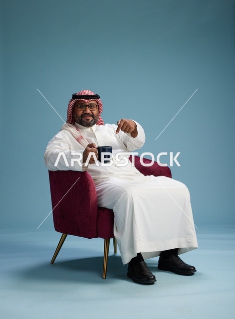 A portrait of an Arab Saudi Gulf man in a white thobe sitting holding a cup of coffee, enjoying hot beverages, with a blue background.