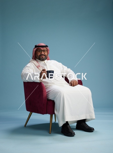بورتريه لرجل عربي سعودي خليجي بثوب أبيض جالس ممسكاً بكوب قهوة, الاستمتاع بالمشروبات الساخنه, خلفيه زرقاء