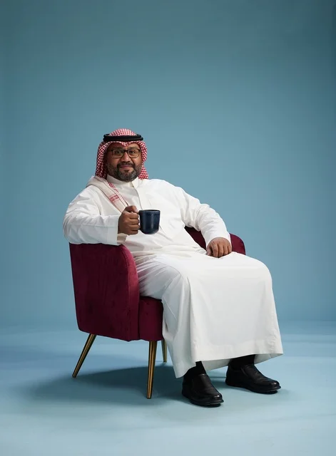 A portrait of an Arab Saudi Gulf man in a white thobe sitting holding a cup of coffee, enjoying hot beverages, with a blue background.