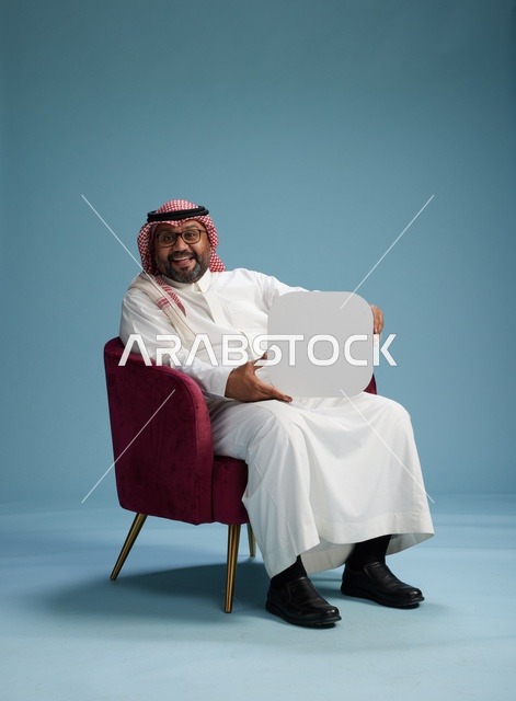 بورتريه لرجل عربي سعودي  خليجي بالزي التقليدي يحمل لافتة مربعة فارغة أثناء جلوسه على كرسي مريح, لوحة اعلانيه بيضاء , خلفيه زرقاء