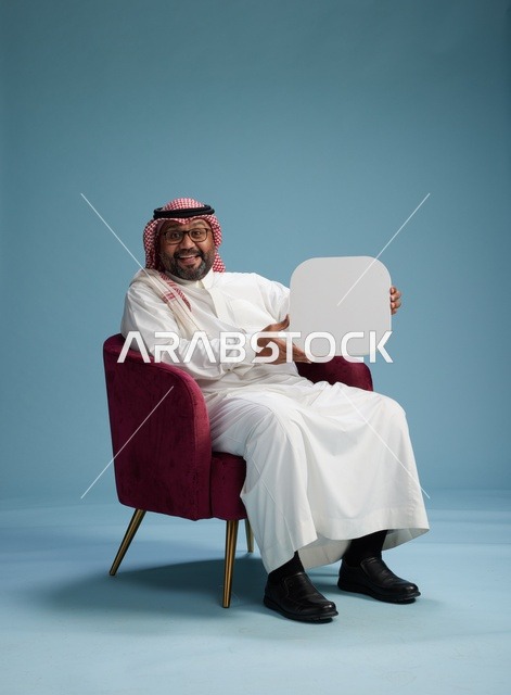 بورتريه لرجل عربي سعودي  خليجي بالزي التقليدي يحمل لافتة مربعة فارغة أثناء جلوسه على كرسي مريح, لوحة اعلانيه بيضاء , خلفيه زرقاء