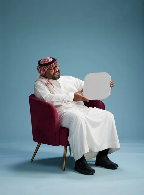A portrait of a Saudi Arabian Gulf man in traditional attire holding a blank square sign while sitting on a comfortable chair, a white billboard, blue background.