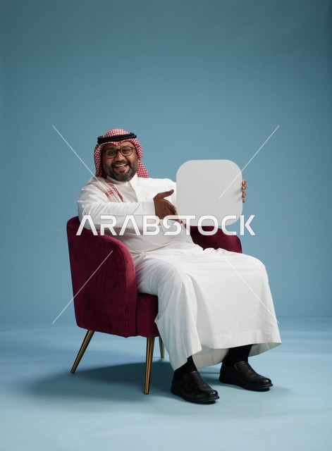 بورتريه لرجل عربي سعودي  خليجي بالزي التقليدي يحمل لافتة مربعة فارغة أثناء جلوسه على كرسي مريح, لوحة اعلانيه بيضاء , خلفيه زرقاء
