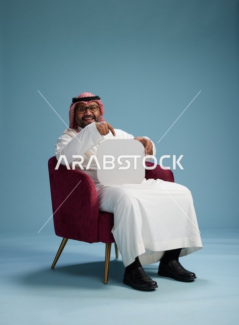 بورتريه لرجل عربي سعودي  خليجي بالزي التقليدي يحمل لافتة مربعة فارغة أثناء جلوسه على كرسي مريح, لوحة اعلانيه بيضاء , خلفيه زرقاء