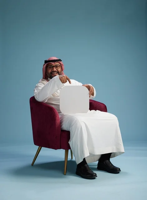 A portrait of a Saudi Arabian Gulf man in traditional attire holding a blank square sign while sitting on a comfortable chair, a white billboard, blue background.