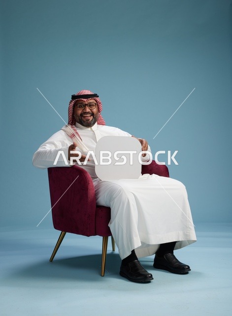 بورتريه لرجل عربي سعودي  خليجي بالزي التقليدي يحمل لافتة مربعة فارغة أثناء جلوسه على كرسي مريح, لوحة اعلانيه بيضاء , خلفيه زرقاء