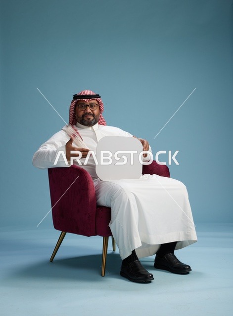بورتريه لرجل عربي سعودي  خليجي بالزي التقليدي يحمل لافتة مربعة فارغة أثناء جلوسه على كرسي مريح, لوحة اعلانيه بيضاء , خلفيه زرقاء