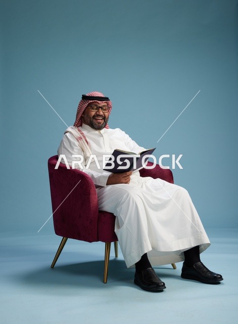 بورتريه لرجل عربي  سعودي  خليجي بثوب أبيض يقرأ كتاباً أثناء جلوسه ويبتسم, الاستمتاع بالقراءه والمطالعه, خلفيه زرقاء