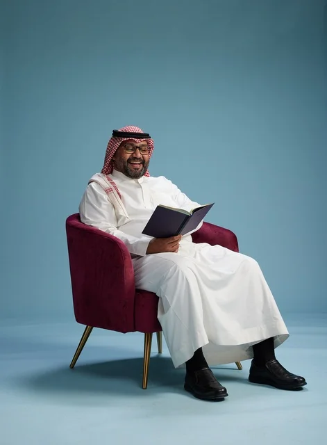 A portrait of an Arab Saudi Gulf man in a white thobe reading a book while sitting and smiling, enjoying reading and studying, with a blue background.