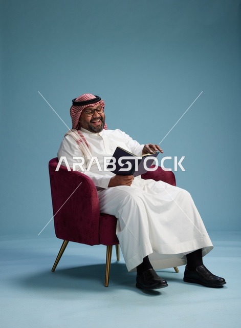بورتريه لرجل عربي  سعودي  خليجي بثوب أبيض يقرأ كتاباً أثناء جلوسه ويبتسم, الاستمتاع بالقراءه والمطالعه, خلفيه زرقاء