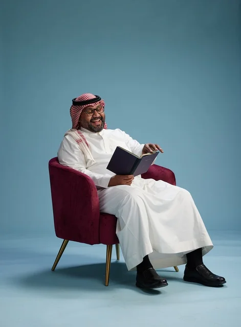 A portrait of an Arab Saudi Gulf man in a white thobe reading a book while sitting and smiling, enjoying reading and studying, with a blue background.