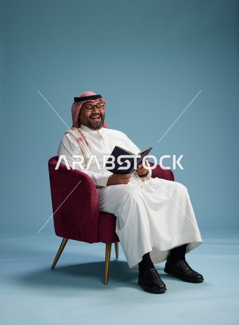 بورتريه لرجل عربي  سعودي  خليجي بثوب أبيض يقرأ كتاباً أثناء جلوسه ويبتسم, الاستمتاع بالقراءه والمطالعه, خلفيه زرقاء
