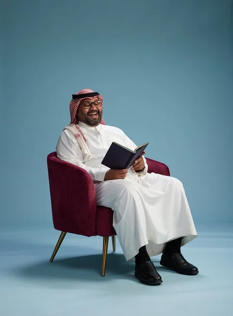 A portrait of an Arab Saudi Gulf man in a white thobe reading a book while sitting and smiling, enjoying reading and studying, with a blue background.