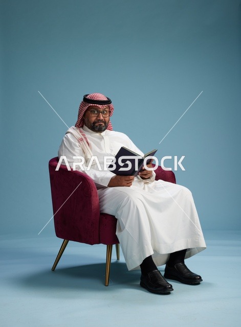 بورتريه لرجل عربي  سعودي  خليجي بثوب أبيض يقرأ كتاباً أثناء جلوسه ويبتسم, الاستمتاع بالقراءه والمطالعه, خلفيه زرقاء