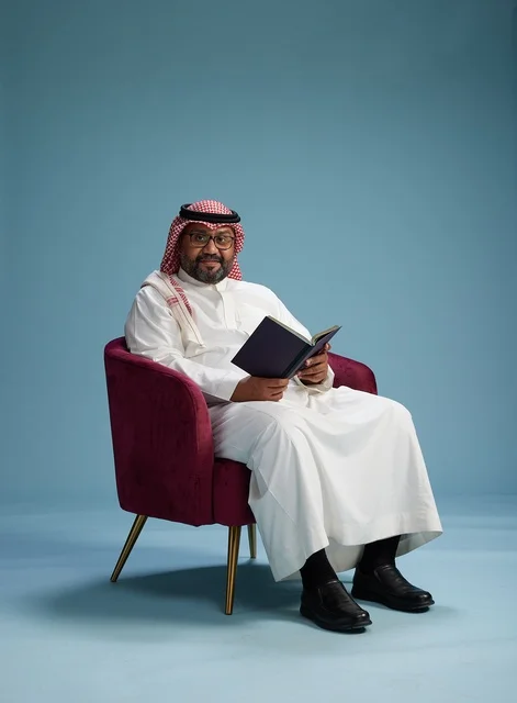 A portrait of an Arab Saudi Gulf man in a white thobe reading a book while sitting and smiling, enjoying reading and studying, with a blue background.