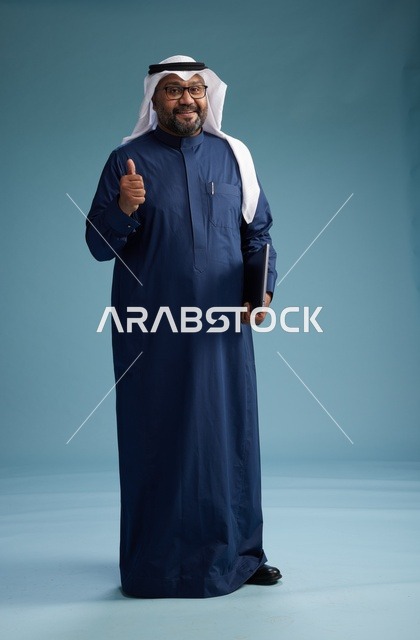 بورتريه لرجل سعودي بثوب أزرق وغترة بيضاء يستخدم الكمبيوتر المحمول, خلفيه زرقاء