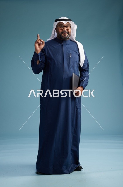 بورتريه لرجل سعودي بثوب أزرق وغترة بيضاء يستخدم الكمبيوتر المحمول, خلفيه زرقاء