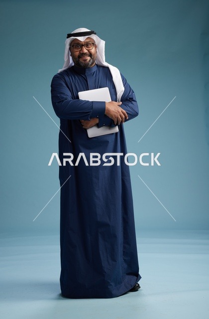 بورتريه لرجل سعودي بثوب أزرق وغترة بيضاء يستخدم الكمبيوتر المحمول, خلفيه زرقاء