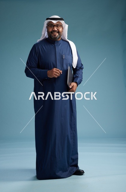A portrait of a Saudi man in a blue thobe and white ghutrah using a laptop, with a blue background.