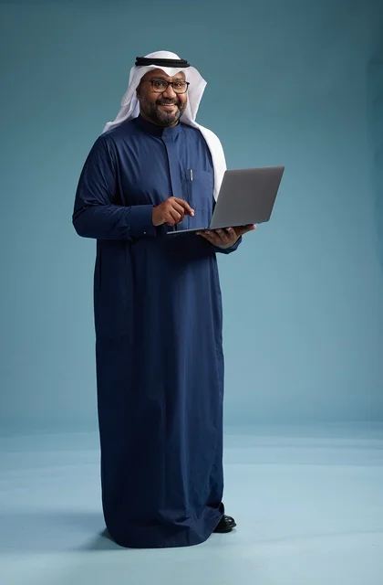 A portrait of a Saudi man in a blue thobe and white ghutrah using a laptop, with a blue background.
