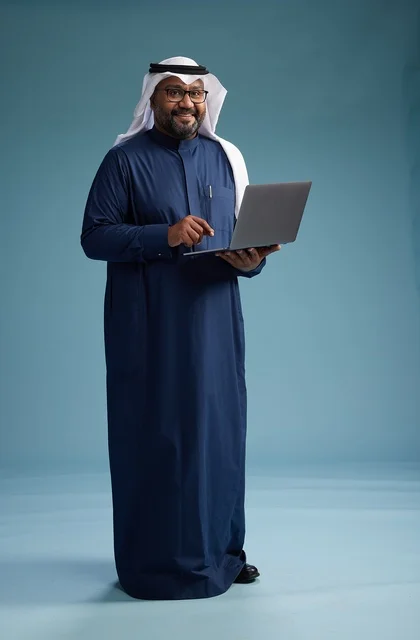 A portrait of a Saudi man in a blue thobe and white ghutrah using a laptop, with a blue background.