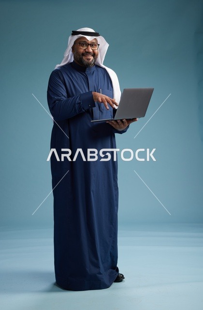بورتريه لرجل سعودي بثوب أزرق وغترة بيضاء يستخدم الكمبيوتر المحمول, خلفيه زرقاء