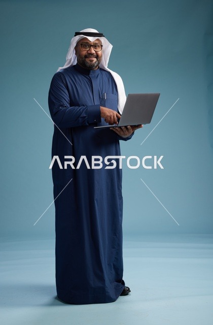 بورتريه لرجل سعودي بثوب أزرق وغترة بيضاء يستخدم الكمبيوتر المحمول, خلفيه زرقاء