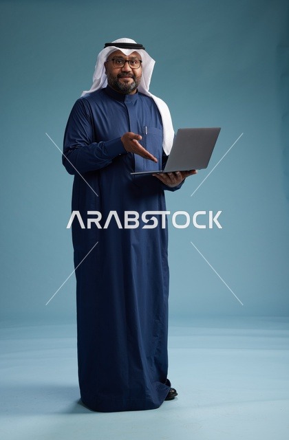 بورتريه لرجل سعودي بثوب أزرق وغترة بيضاء يستخدم الكمبيوتر المحمول, خلفيه زرقاء