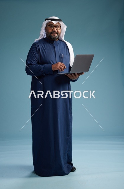 بورتريه لرجل سعودي بثوب أزرق وغترة بيضاء يستخدم الكمبيوتر المحمول, خلفيه زرقاء