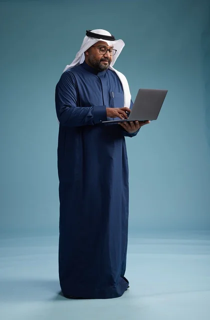 A portrait of a Saudi man in a blue thobe and white ghutrah using a laptop, with a blue background.